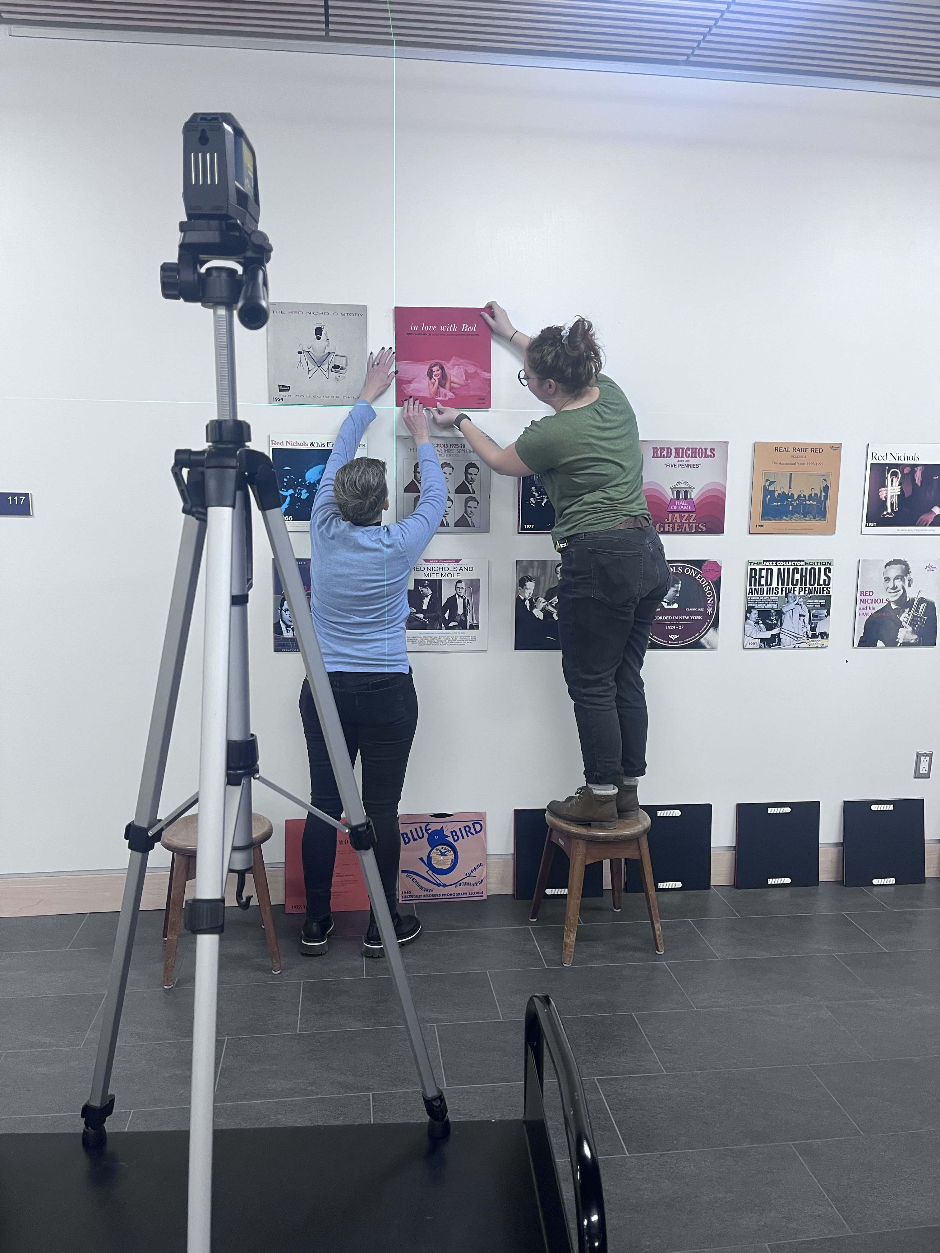 Marina Kenner and Lorrie Rands mount exhibit materials to the wall.