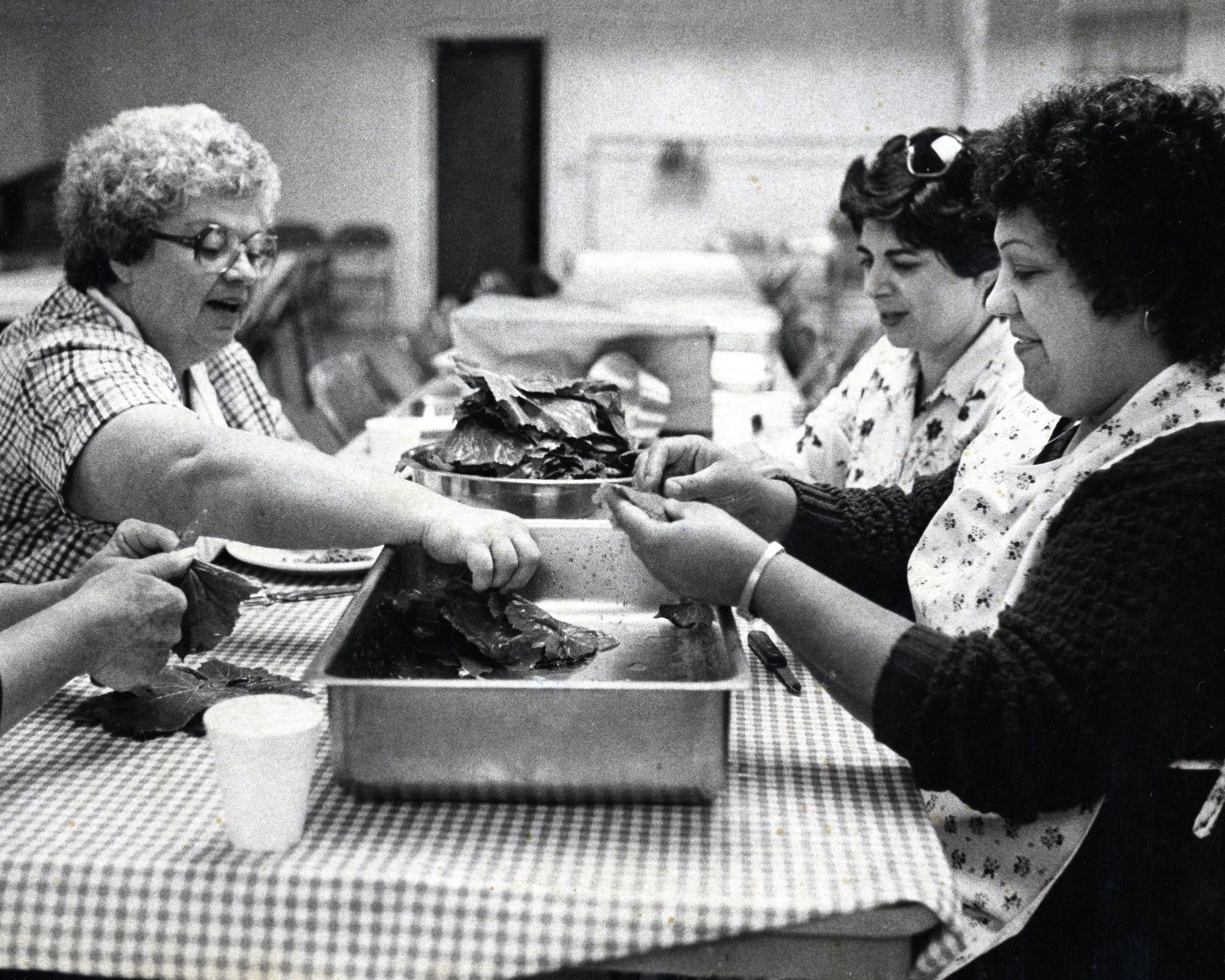 Three women sitting at a table preparing food using grape leaves.